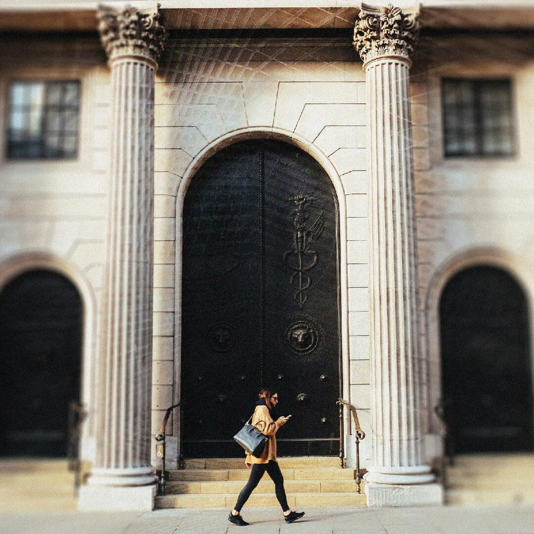 Person walking past a grand building with tall columns and ornate black doors, holding a phone and wearing a coat and bag.