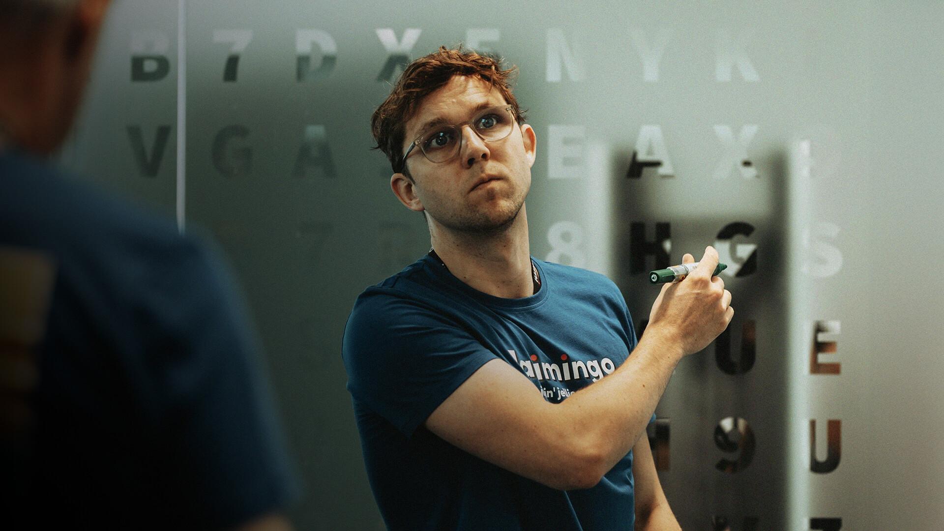 A person with glasses writes on a transparent board covered in letters and numbers, wearing a blue shirt, focused on the task.