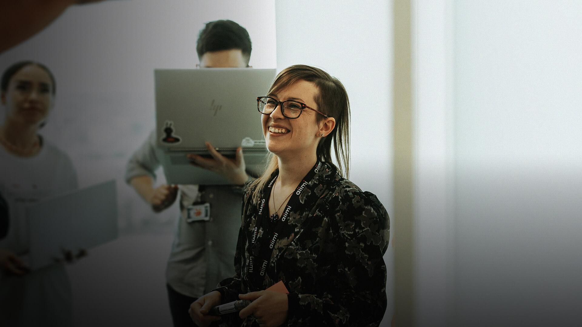 A person smiling, wearing glasses and a patterned shirt, stands in a bright room with others holding laptops in the background.