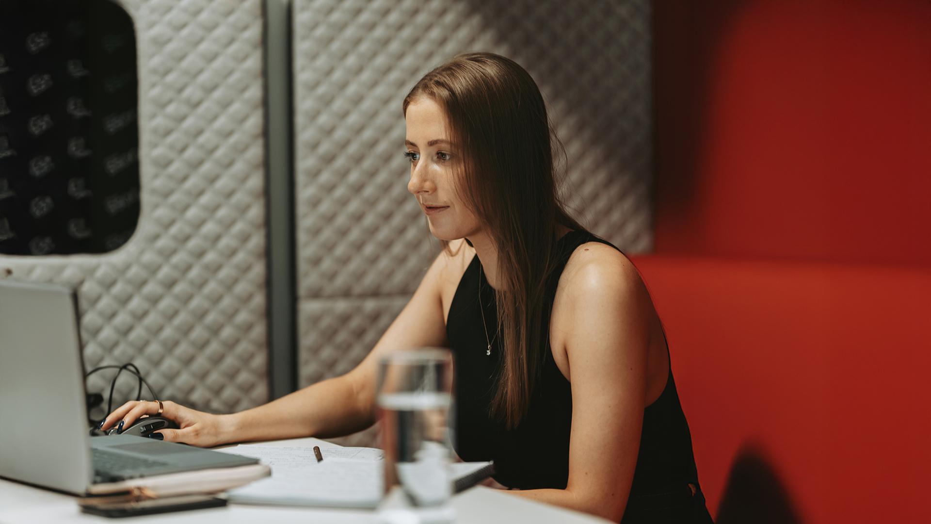 Woman working on a laptop in a modern office space, with a notebook and a glass of water on the table.