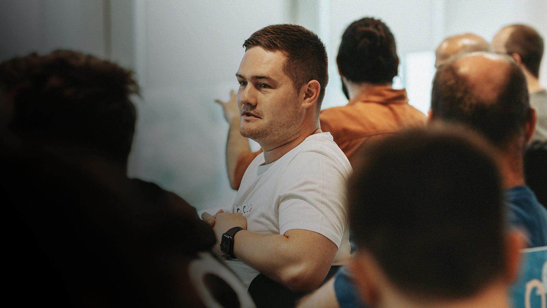 A man in a white shirt sits attentively in a crowded room, facing forward. Others around him are engaged in the same direction.