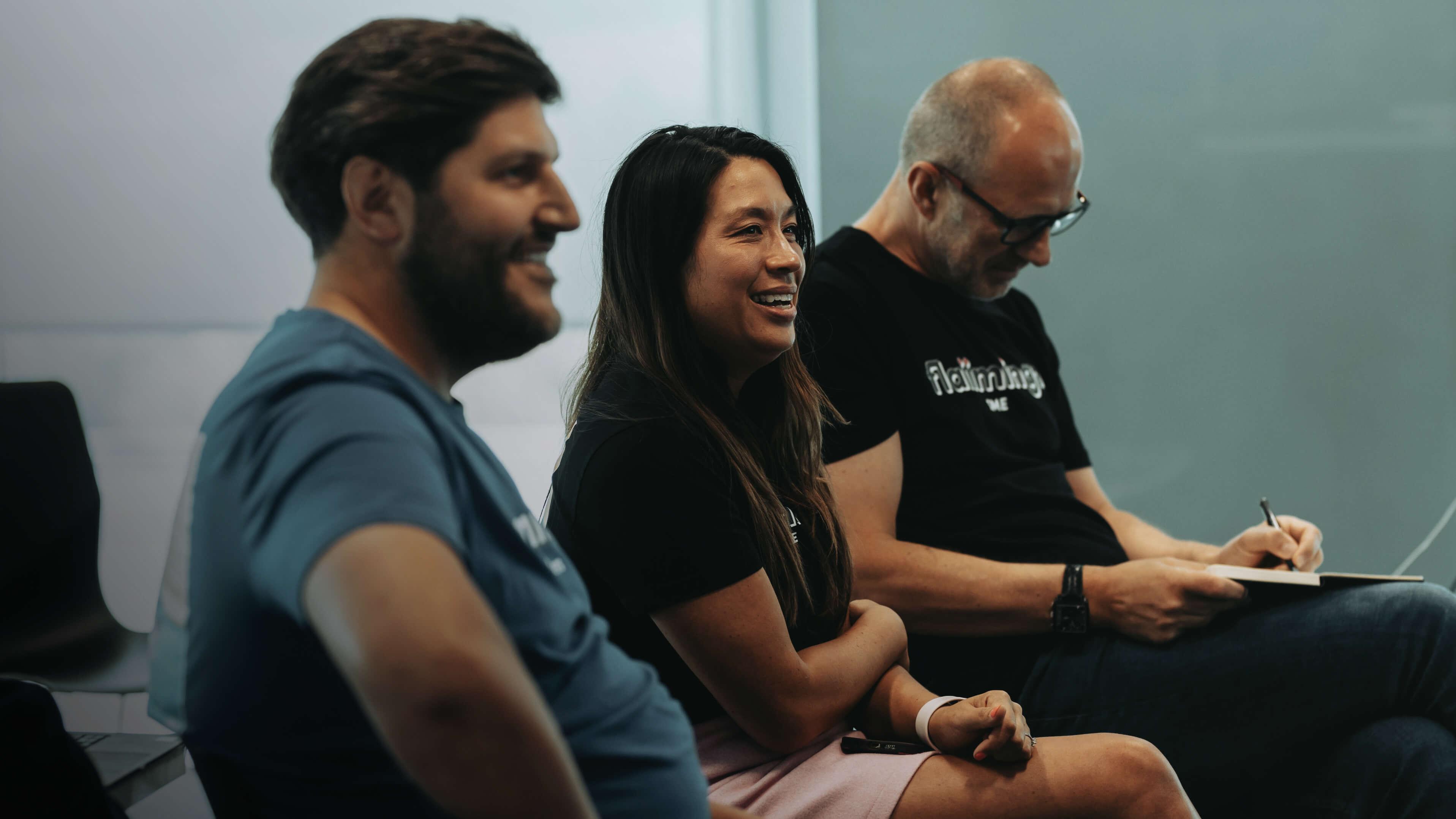 Three people sitting and smiling in a casual setting, with one person writing in a notebook.