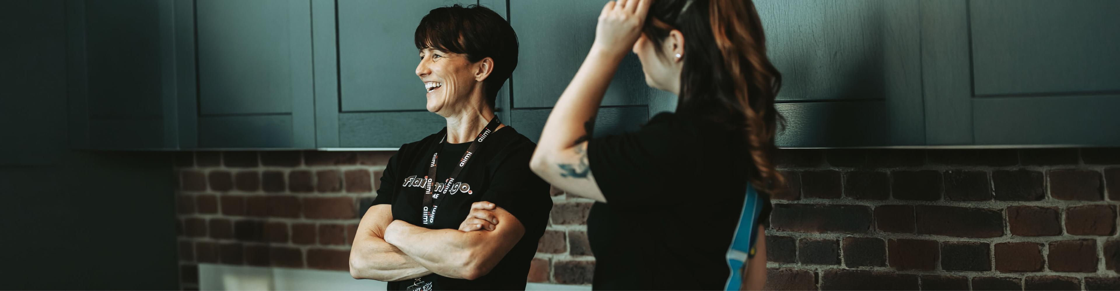 Two people wearing black shirts chatting and smiling in a kitchen with brick walls and dark cabinets.