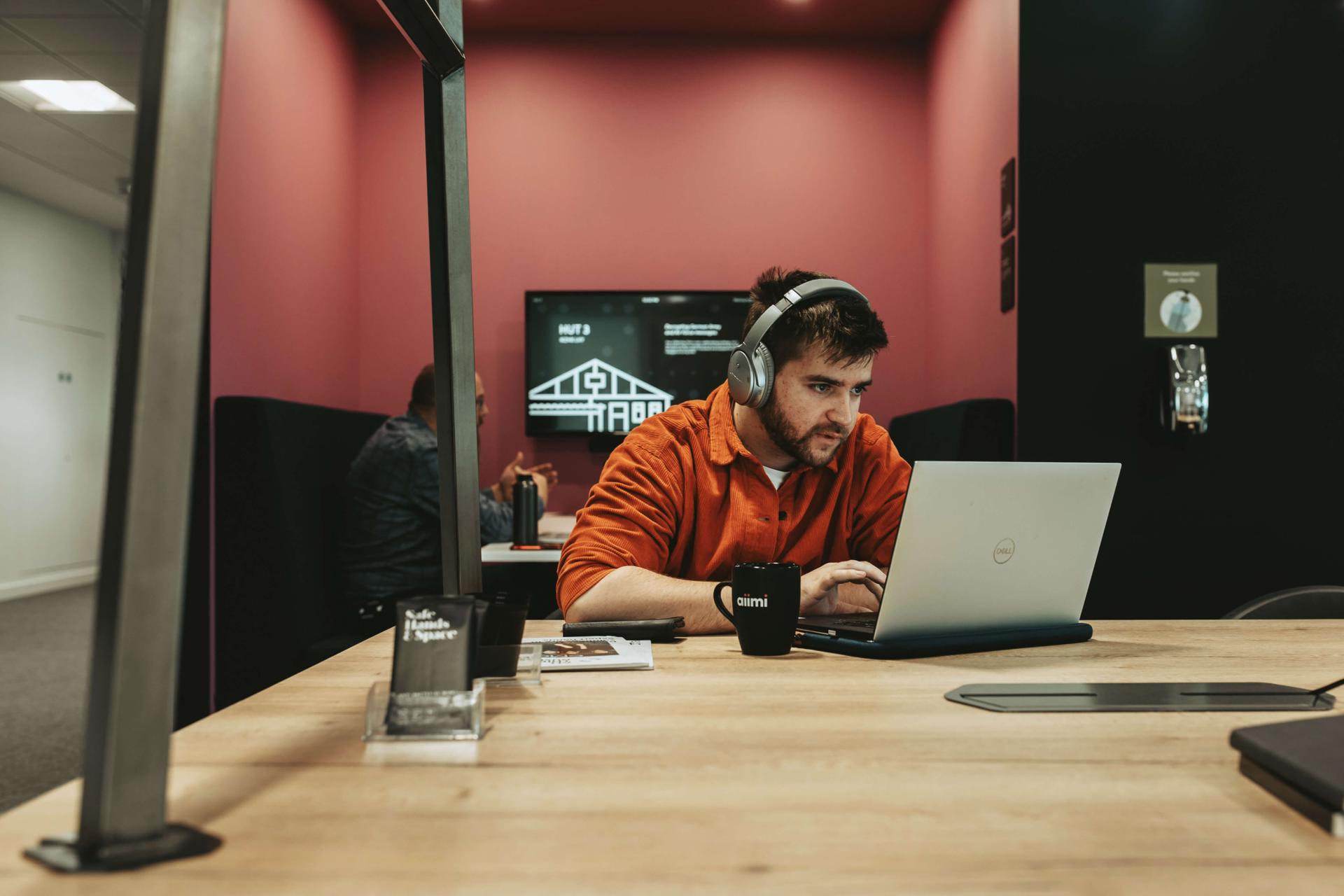 Man in an orange shirt and headphones works on a laptop at a wooden table, with a black mug and brochures nearby, in a modern office space.