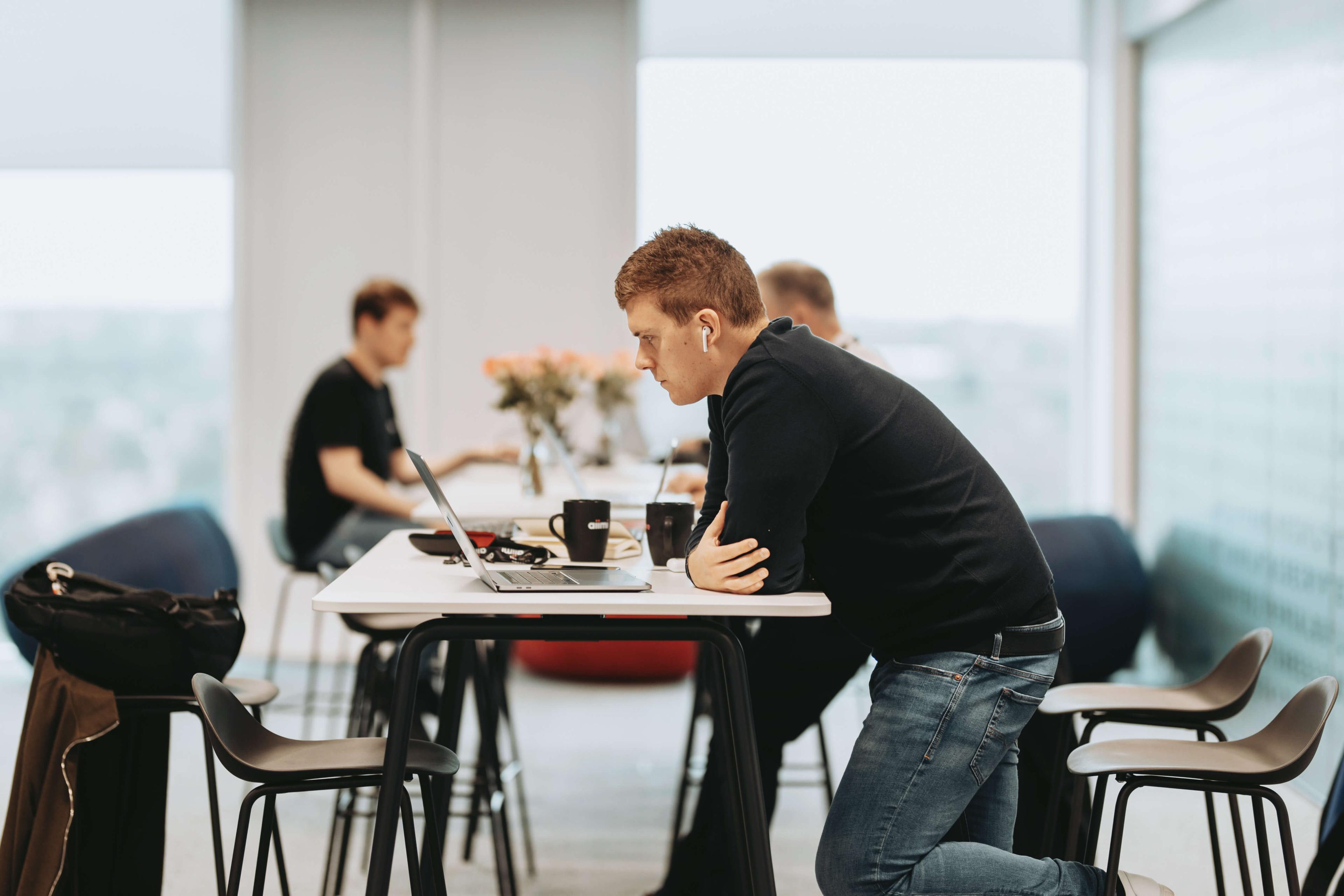 Person in a black sweater leans over a laptop at a high table in a modern office, with two others blurred in the background.