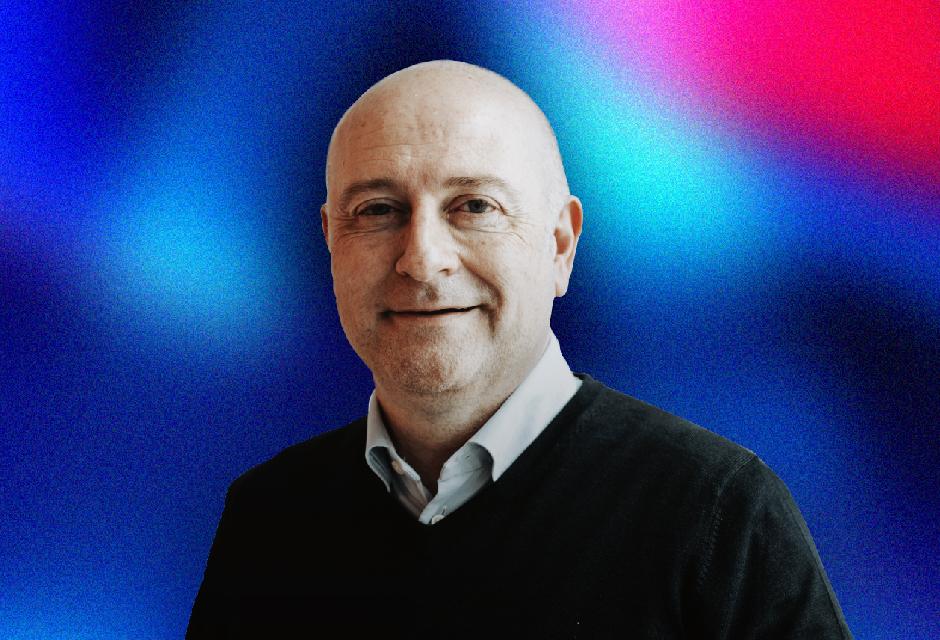 A smiling man with a bald head wearing a dark sweater over a collared shirt, set against a vibrant blue and red gradient background.