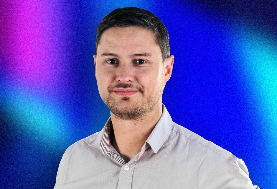 A man in a light shirt stands against a vibrant blue and pink gradient background, looking at the camera.