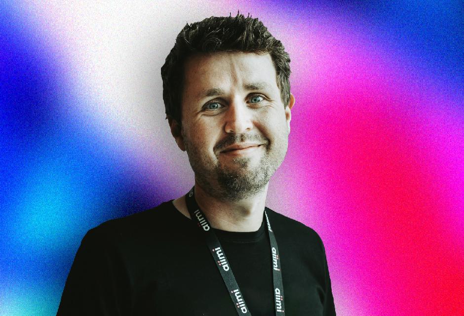 Man smiling, wearing a black shirt and lanyard, standing against a vibrant gradient background of blue, pink, and white.