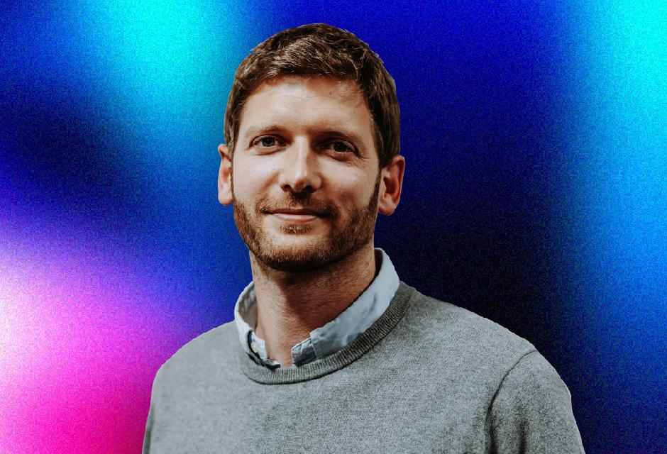 A man with short hair and a beard, wearing a gray sweater over a collared shirt, stands against a vibrant blue and purple gradient background.