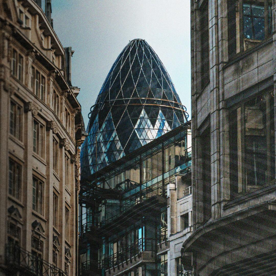 A view of the Gherkin skyscraper in London, framed by surrounding historic buildings, under a clear sky.