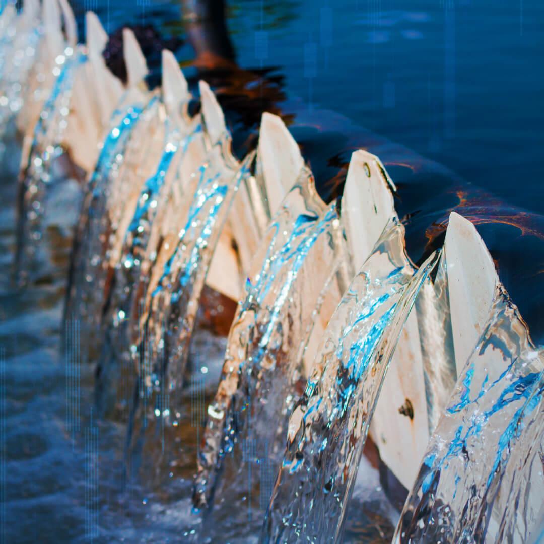 Water cascades over a series of white, triangular barriers into a blue body of water, creating a dynamic, flowing pattern.