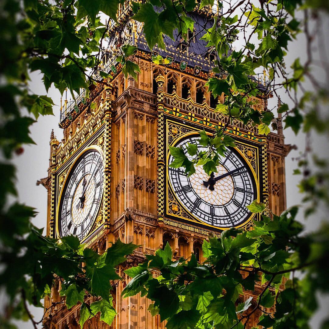 The clock tower of Big Ben framed by green leaves, showcasing intricate details and a clear clock face against a cloudy sky.