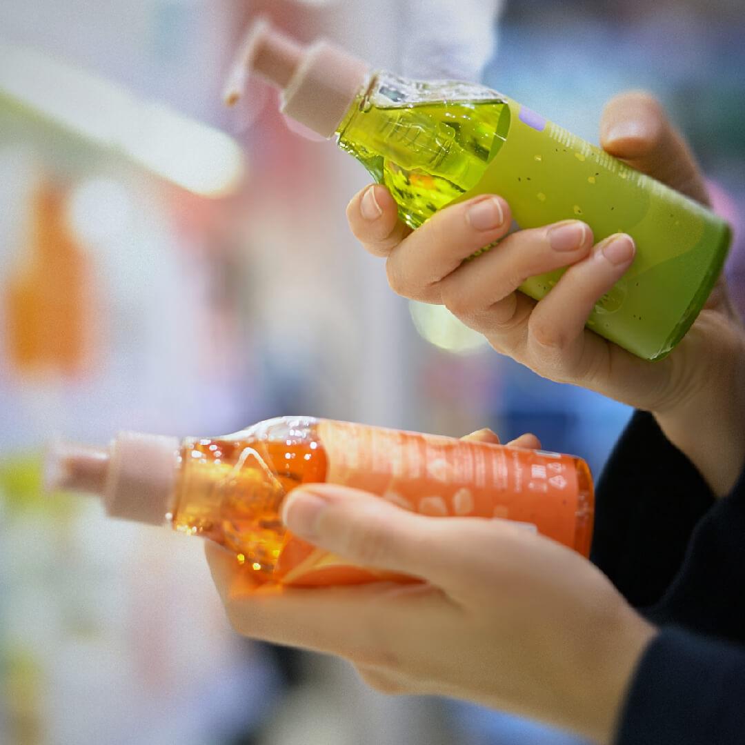 Two hands holding pump bottles; one green and one orange. The background is a blurred store aisle.