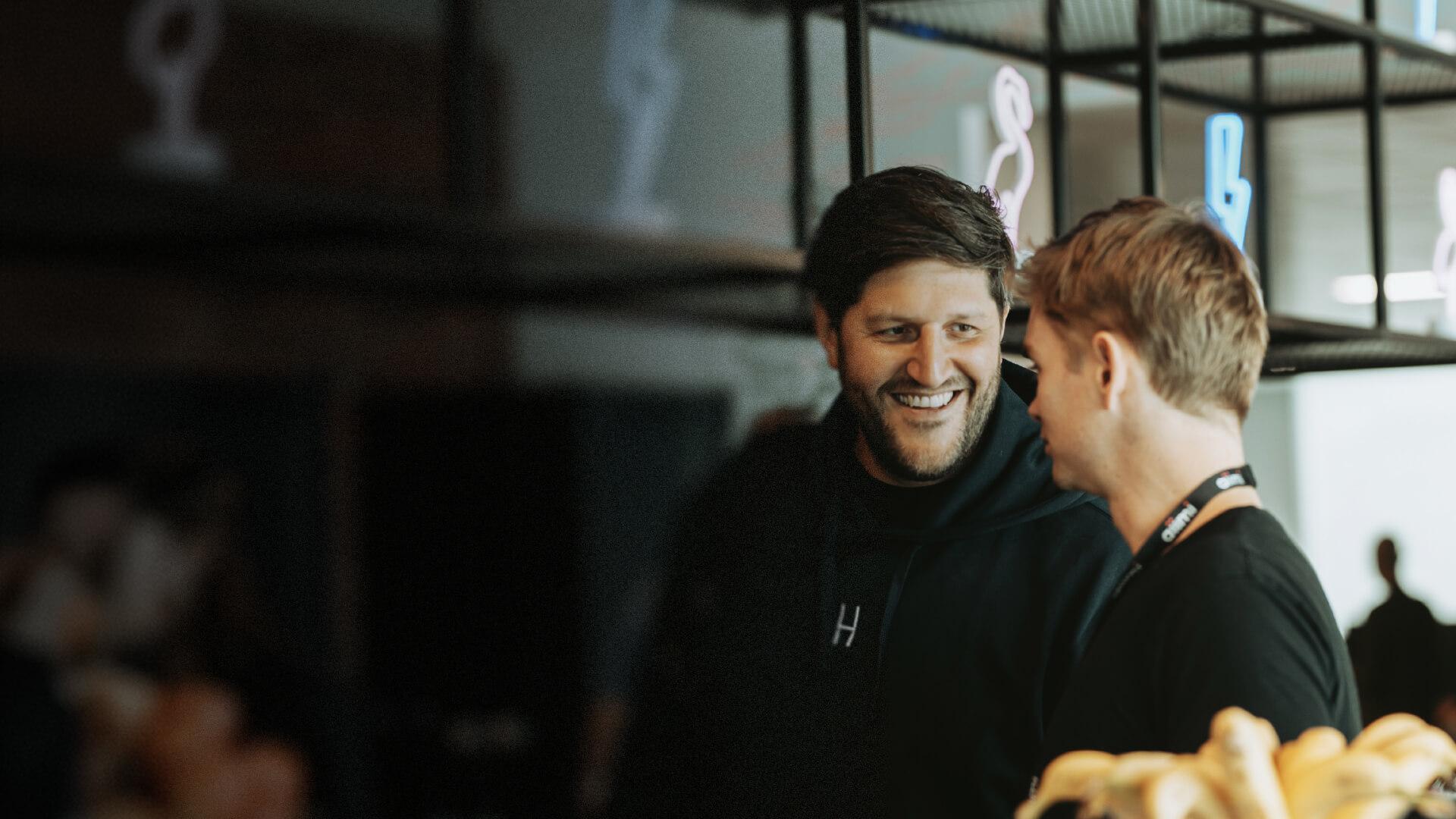 Two men smiling and talking in a bright room with blurred people and neon signs in the background.