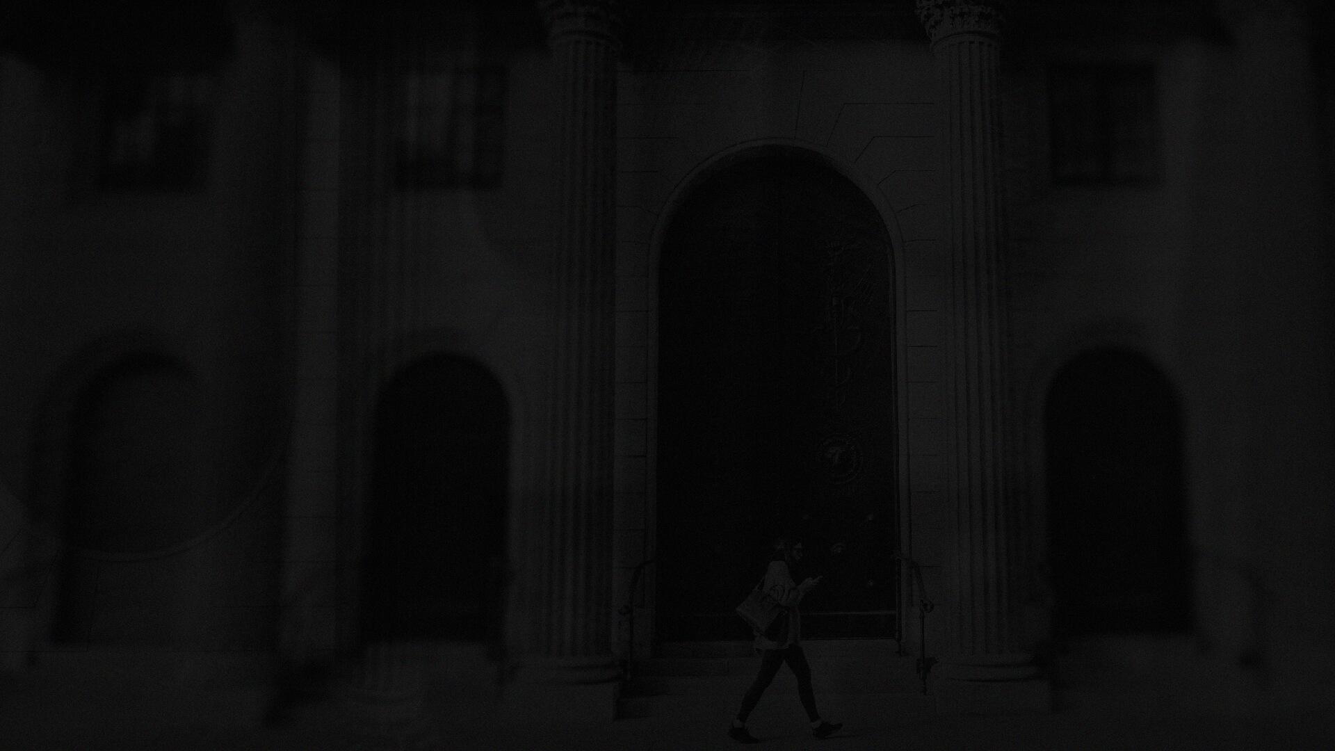 Person walking past a large, arched doorway of a building with tall columns in a dimly lit scene.