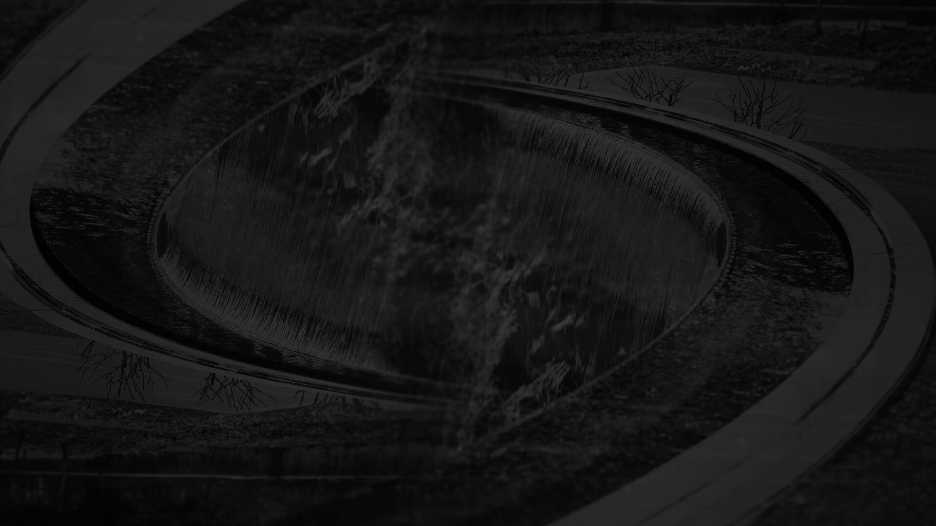 Dark image of an elliptical water feature with flowing water and surrounding curved pathways and sparse plants.