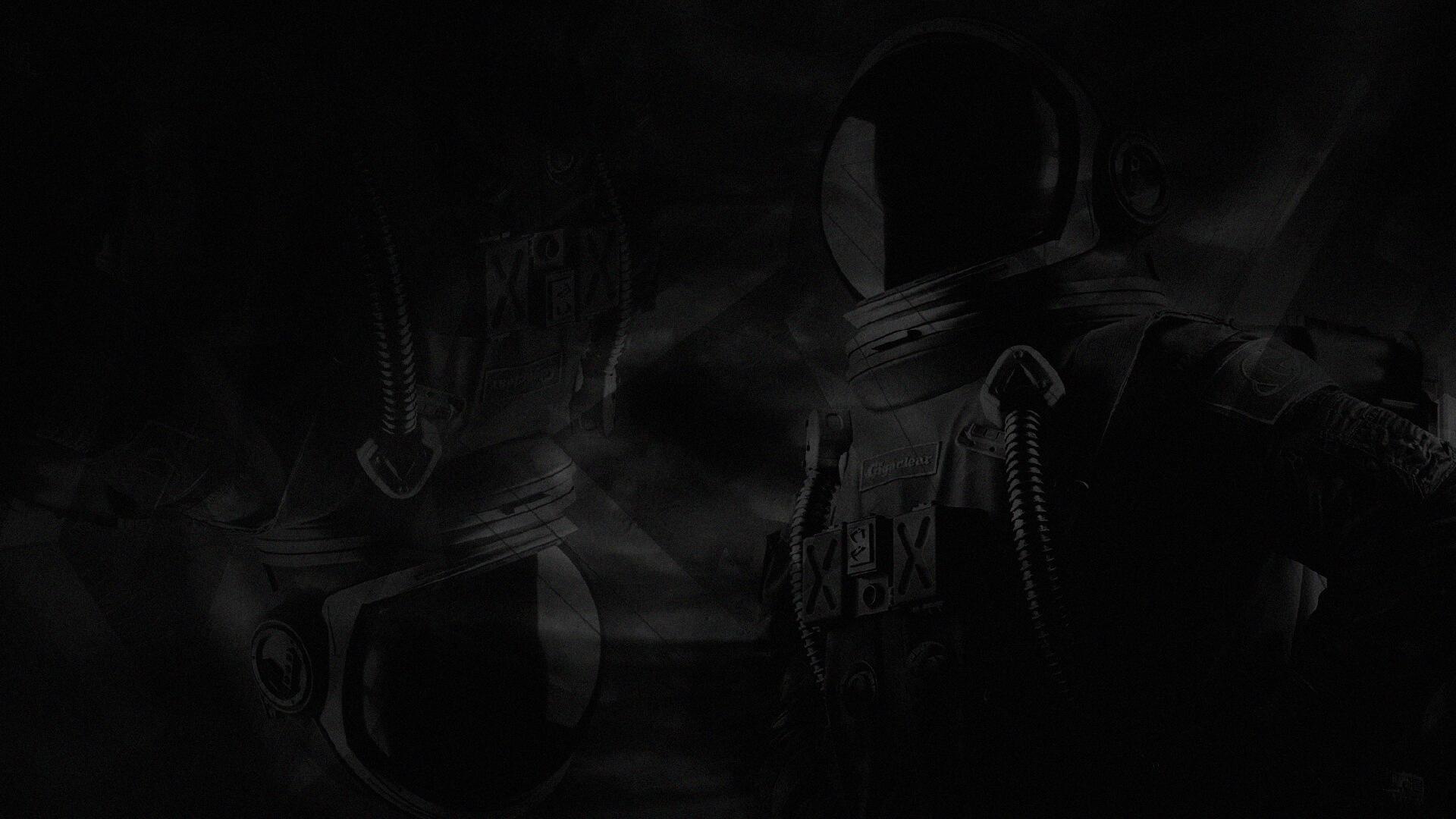 A dark, shadowy image featuring two astronauts in reflective helmets and detailed suits, set against a blurred, indistinct background.