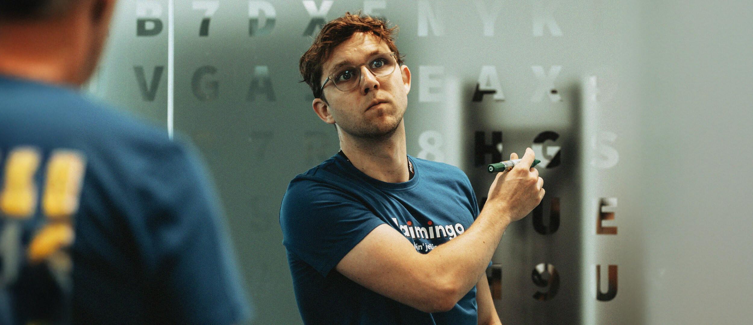 Man in glasses wearing a blue t-shirt holds a marker and points at a frosted glass panel stenciled with letters and numbers.