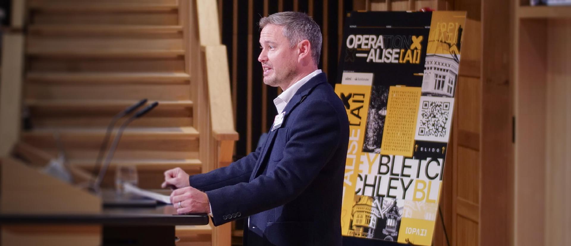 A man speaks at a podium in front of a wooden staircase, with a colorful poster featuring text and architectural images in the background.