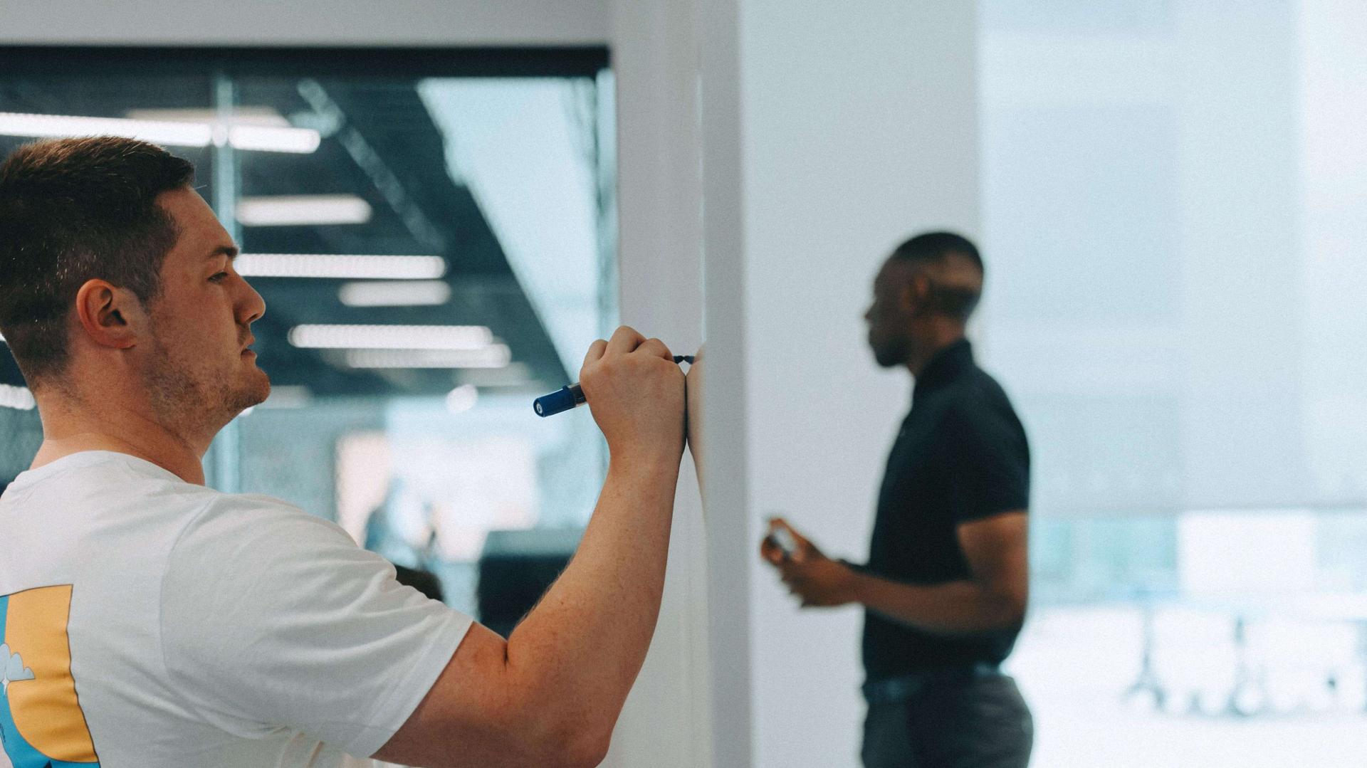Two people in an office: one writes on a whiteboard, the other stands holding a phone, with a modern interior in the background.
