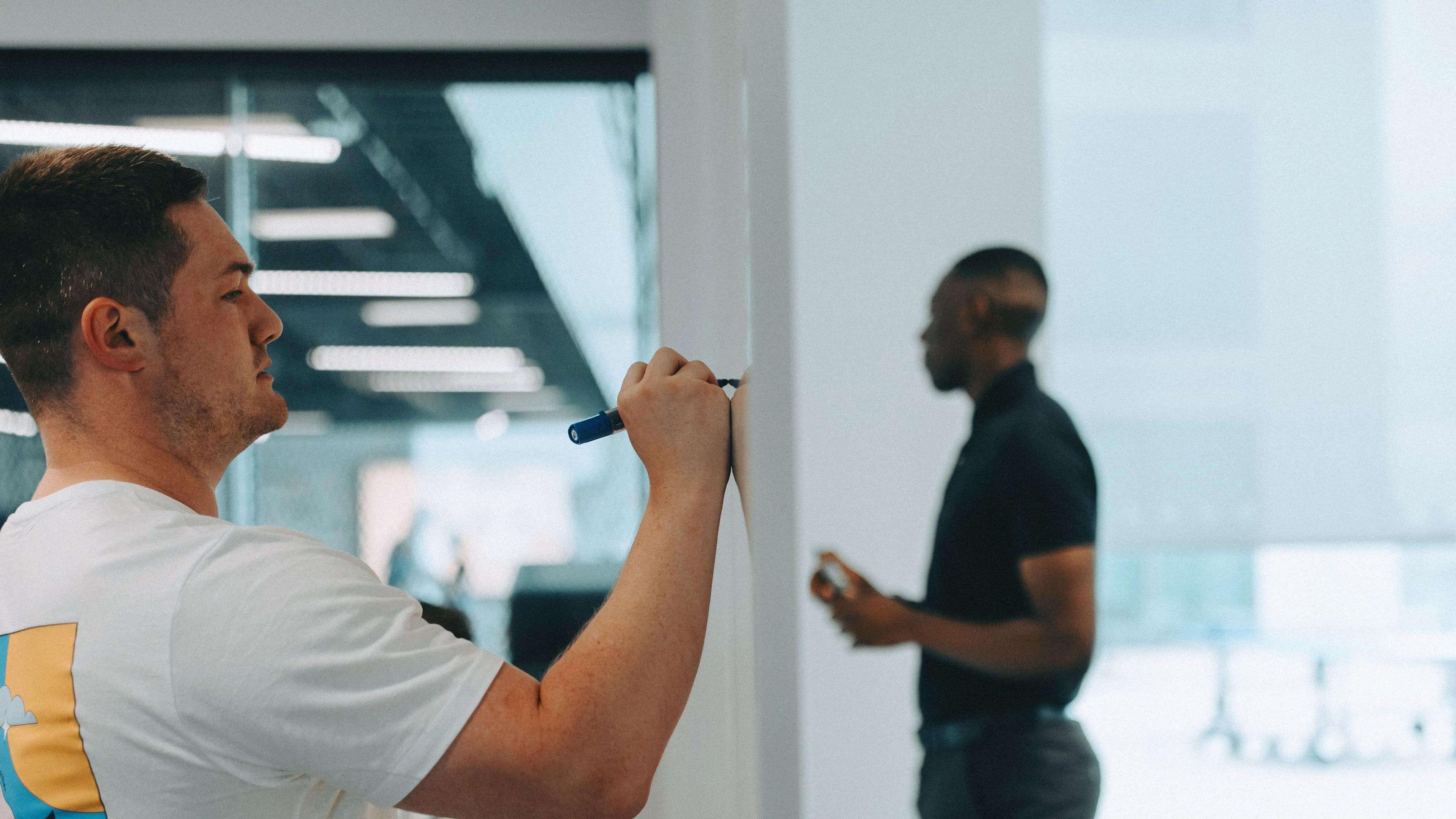 Two people in an office: one writes on a whiteboard, the other stands holding a phone, with a modern interior in the background.